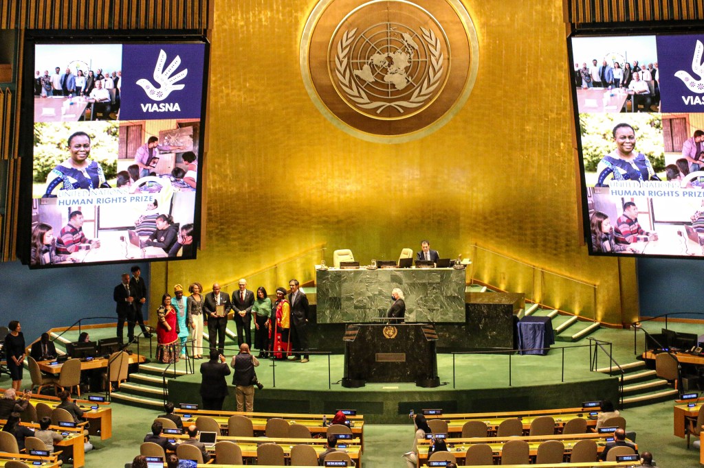A scene from a United Nations event featuring the presentation of the UN Human Rights Prize, with a large screen displaying the Viasna organization and images of individuals involved in human rights advocacy, alongside a group of people on stage.