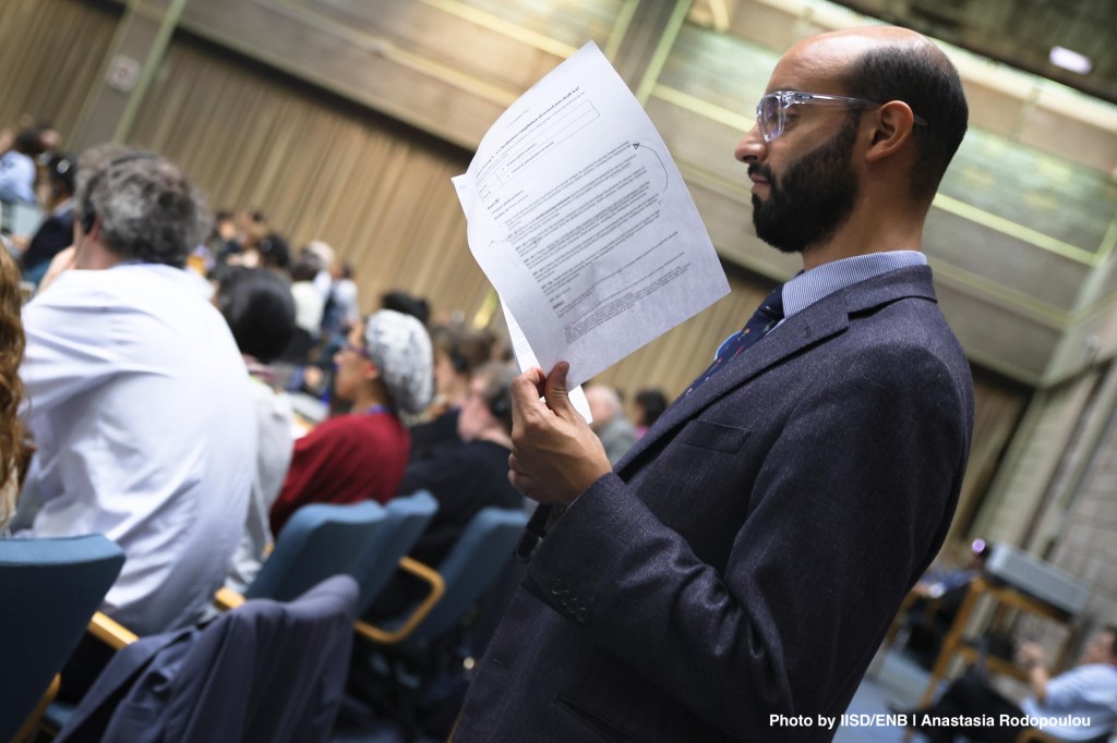 A man in a suit stands reading a document while observing an audience at an event.